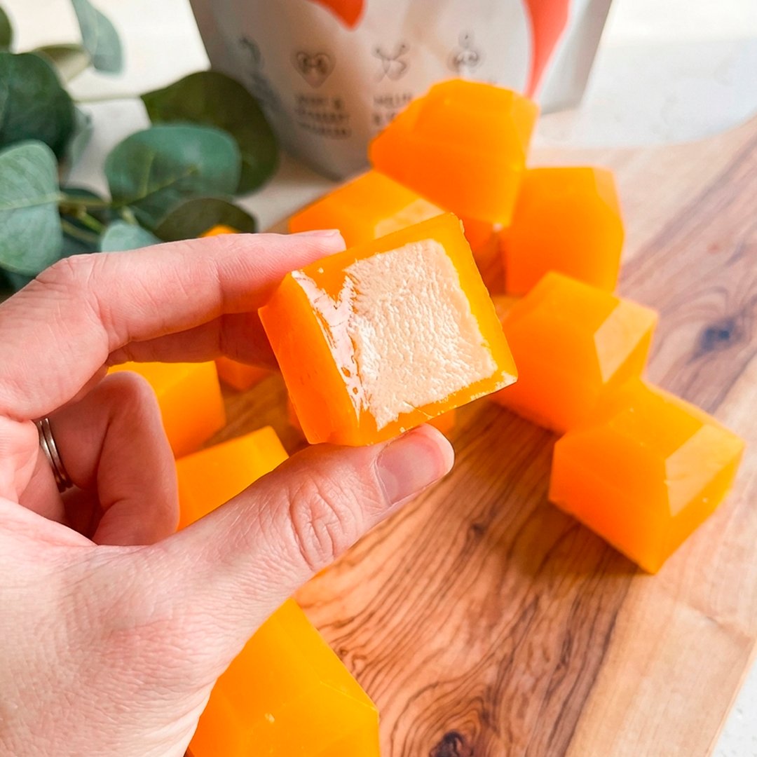 Hand holding a vivid orange mango jelly cube showing cream cheese filling inside, more cubes in background on wooden board